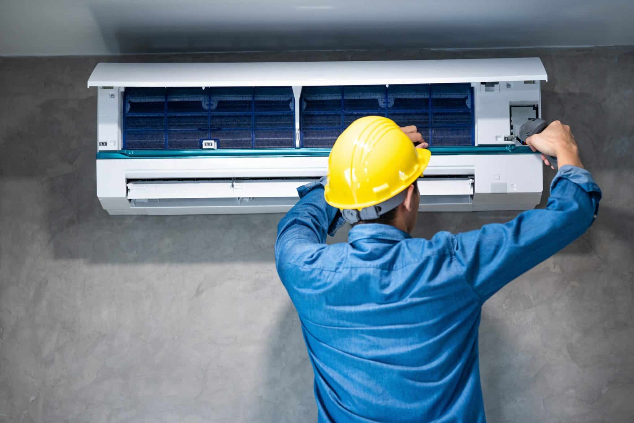 tecnico effettua la manutenzione di un condizionatore a muro, con casco giallo e camicia da lavoro azzurra
